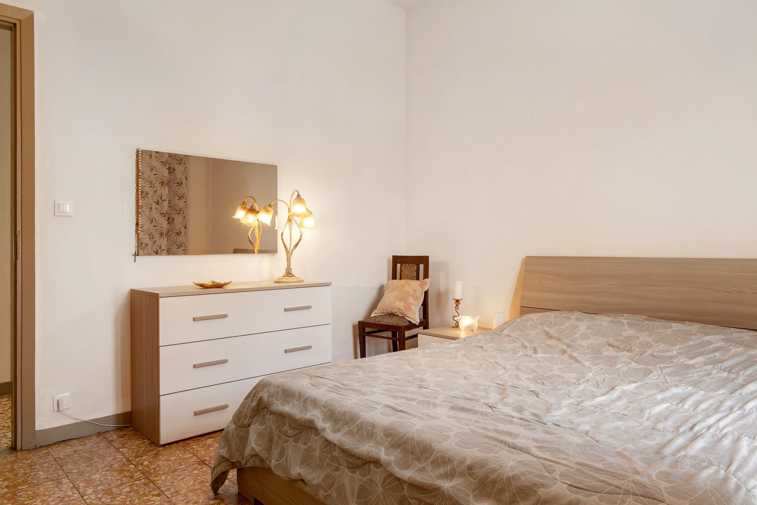 A bedroom with a bed, a dresser with drawers, a chair, a nightstand with a lamp, and a wall-mounted mirror.