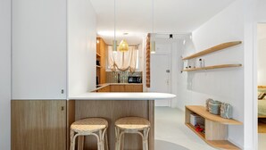 Une cuisine moderne avec un coin bar, deux tabourets en osier, des étagères en bois et un luminaire suspendu.