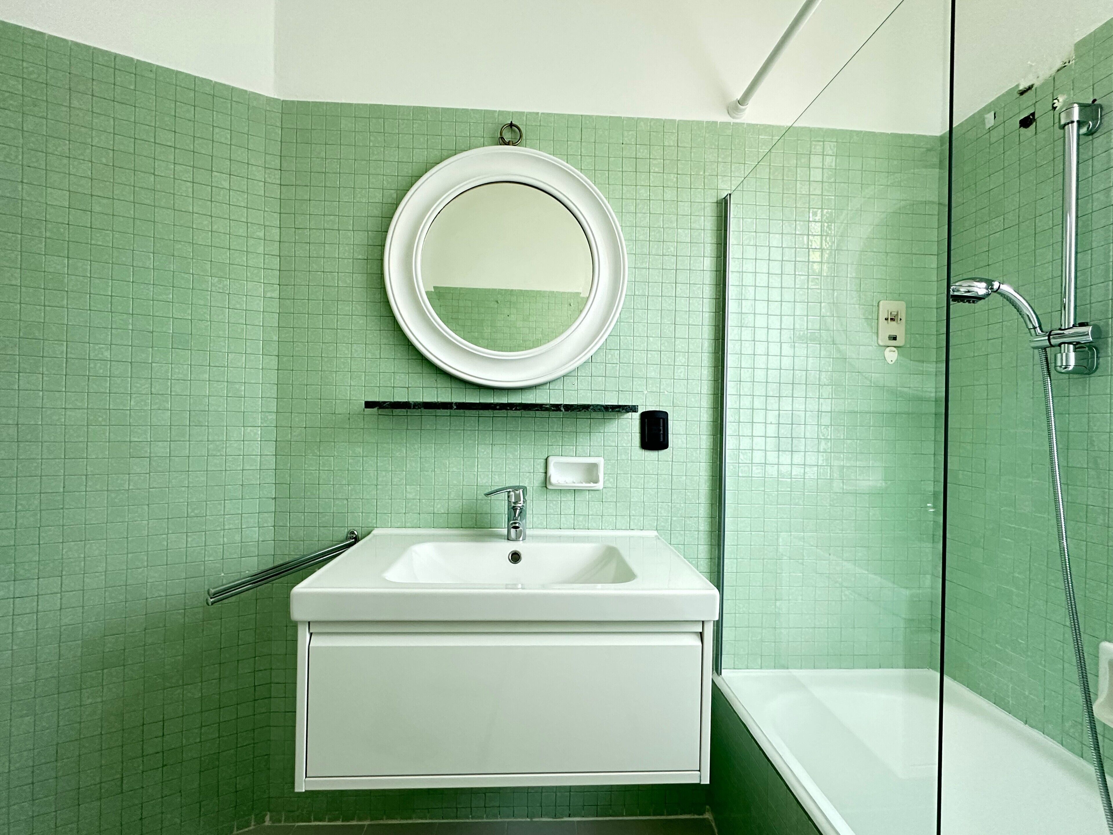 Un bagno con lavandino bianco, uno specchio, una doccia e pareti rivestite di piastrelle verdi.