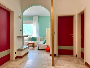 Un pasillo que conduce a una habitación con una puerta roja, un sofá blanco con cojines verdes y una ventana con cortinas blancas.
