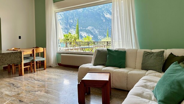 A living room with a sofa, a small table, a dining table with chairs, and a view of mountains through a large window. - Villa 'The Queen Of Garda' with Lake View, Private Garden and Balcony (Riva del Garda)