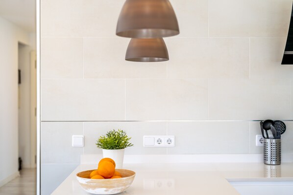 Una cucina moderna con un piano di lavoro bianco, una ciotola di frutta, una pianta in vaso e un lampadario.