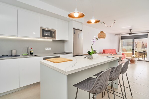 A modern kitchen with white cabinets, a built-in microwave, a stainless steel refrigerator, a marble island with a cutting board, and a vase with a plant.