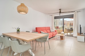 A modern living room with a wooden dining table, grey chairs, a red sofa, and a large window with curtains.