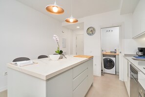 A modern kitchen with a central island, pendant lighting, a washing machine, and a clock on the wall.