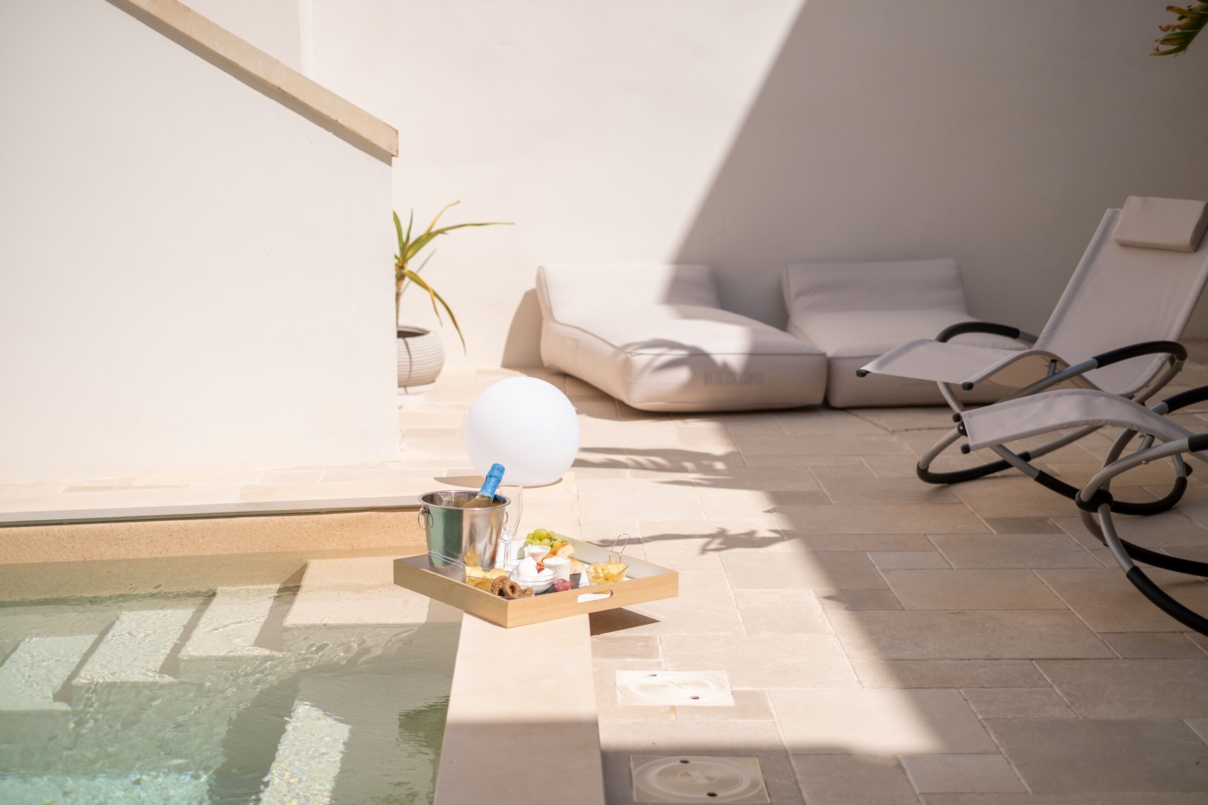 Zona de piscina amb una cadira relaxant, un espai d'seients amb coixins i una bandeja amb menjar i begudes.