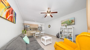 A modern living room with a grey sofa, a yellow armchair, a white coffee table, and a ceiling fan. - Lovely 1BR1BA Condo  ~ Walk to Beach ~ Pool ~ BBQ (Oranjestad)