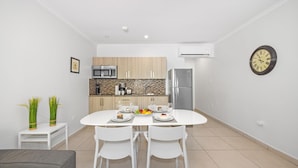 A modern kitchen with white cabinets, a stainless steel refrigerator, and a microwave. There is a dining table with white chairs and a clock on the wall. - Lovely 1BR1BA Condo  ~ Walk to Beach ~ Pool ~ BBQ (Oranjestad)