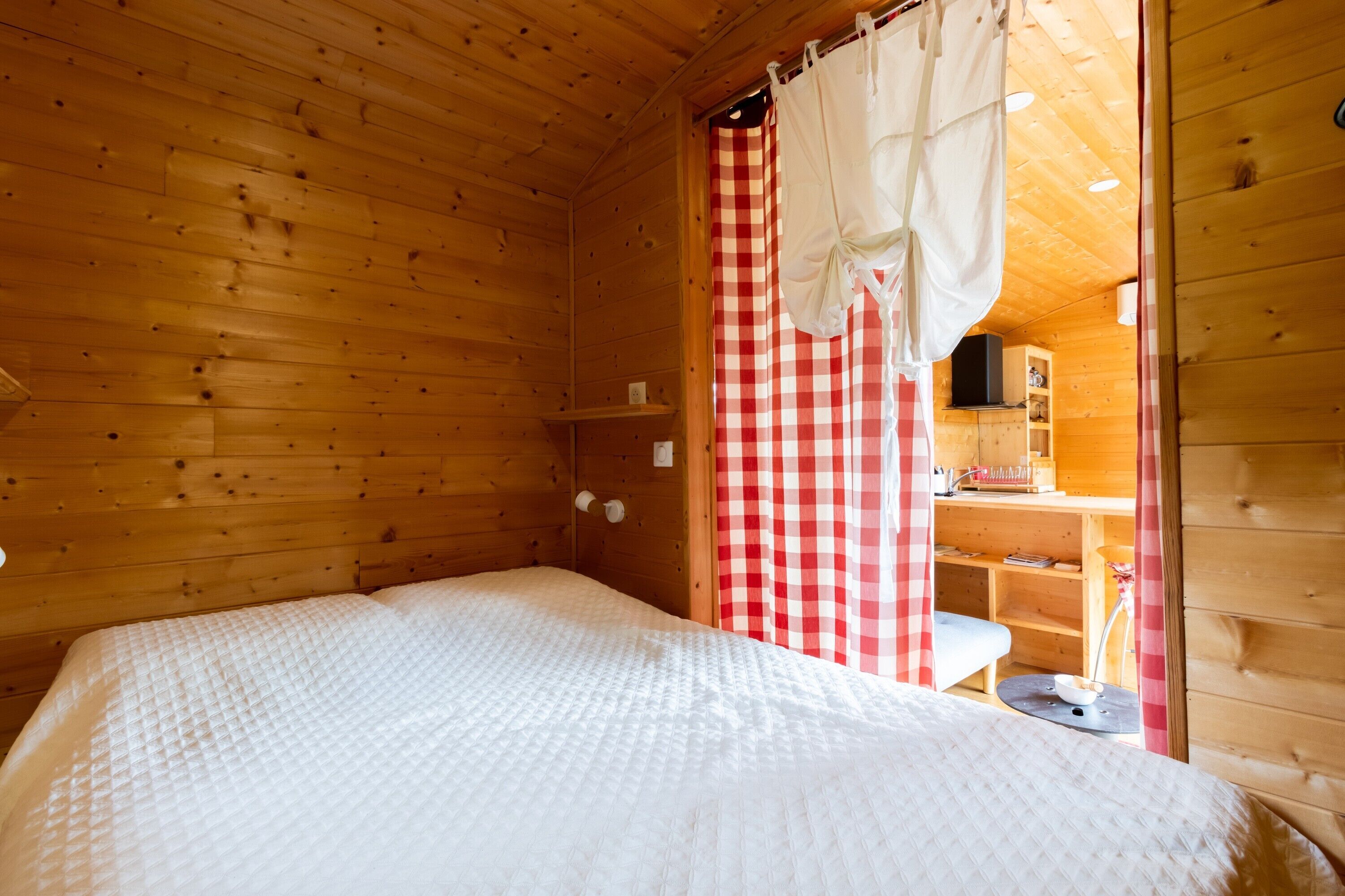 A wooden cabin room with a bed, a checkered curtain, and a small table with a lamp.