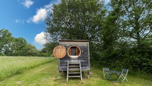 Eine kleine Holzhütte mit rundem Fenster und einer Tür, einer Leiter und zwei Stühlen davor.