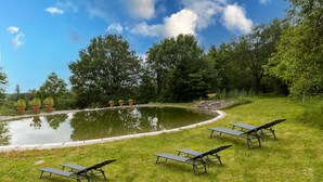 Un étang dont l’eau reflète le paysage environnant, bordé de pelouse et d’arbres, avec des espaces de repos aménagés à l’extérieur.