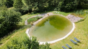 Une vue aérienne d’un étang circulaire entouré de pelouse et d’arbres, avec une promenade dallée et quelques chaises longues.