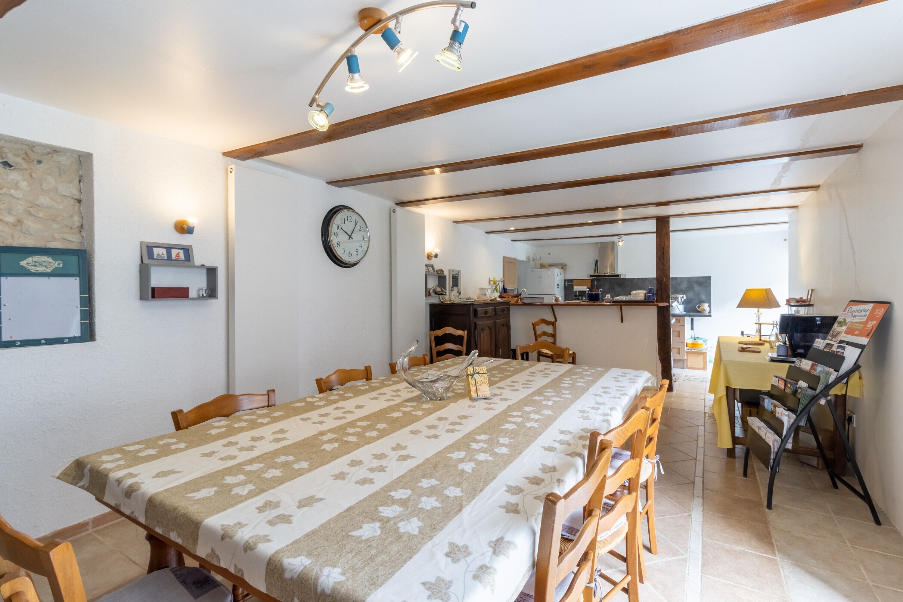 Un espace repas avec une longue table, des chaises, une horloge et une cuisine en arrière-plan.