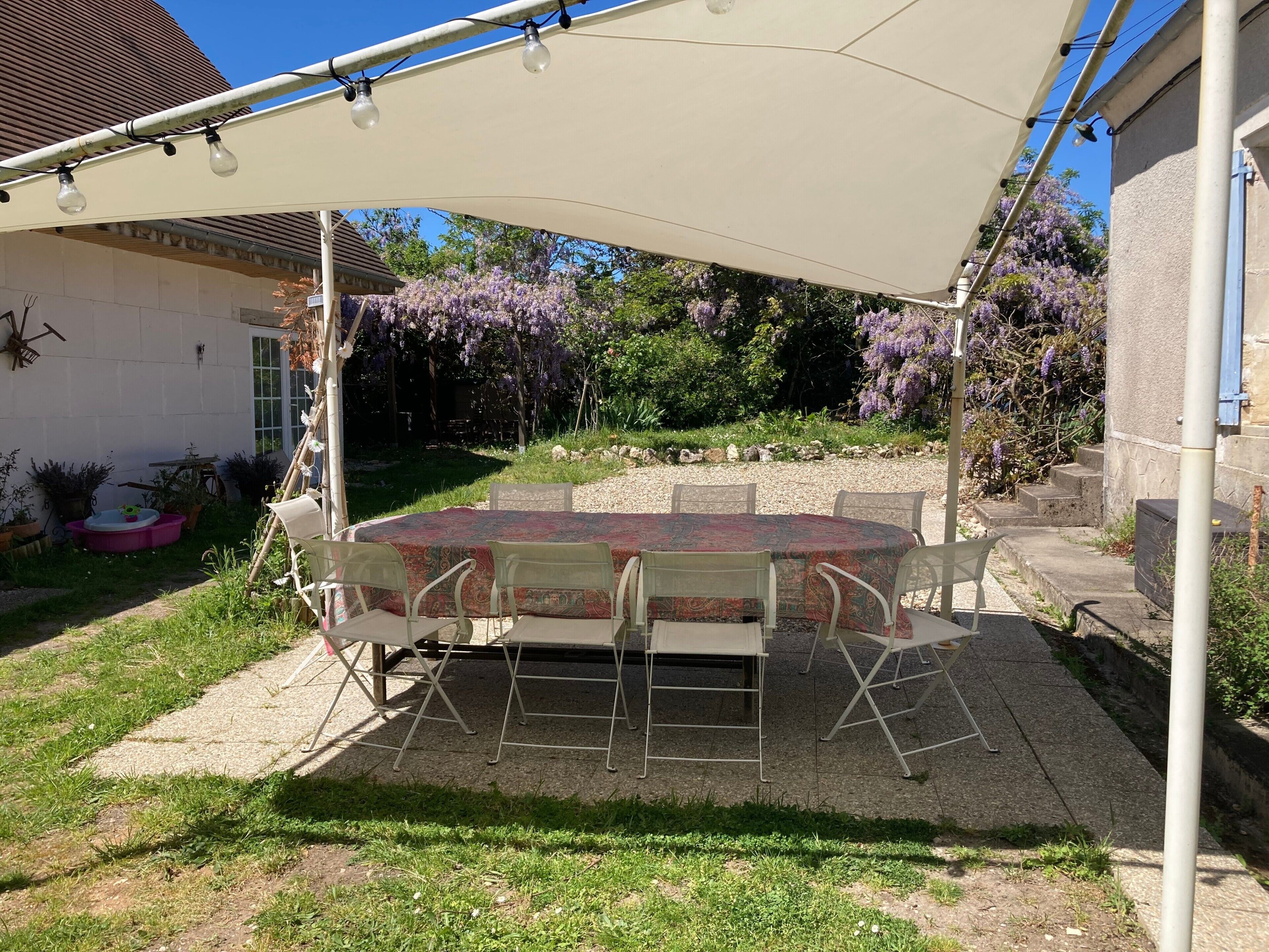 Eine überdachte Terrasse im Freien mit Tisch und Stühlen, umgeben von einem Garten mit violetten Blumen.
