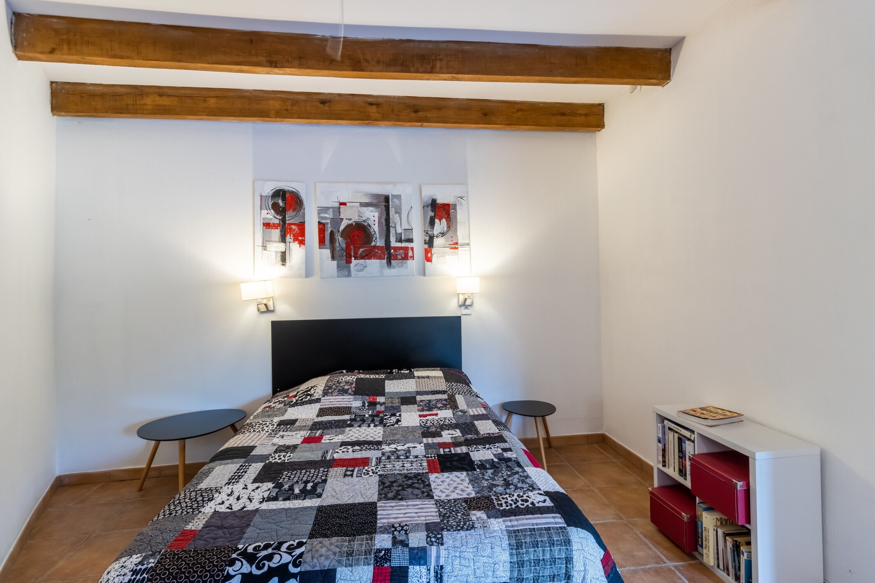 A bedroom with a quilted bedspread, a wooden ceiling beam, a wall-mounted shelf with books, and a small round table.