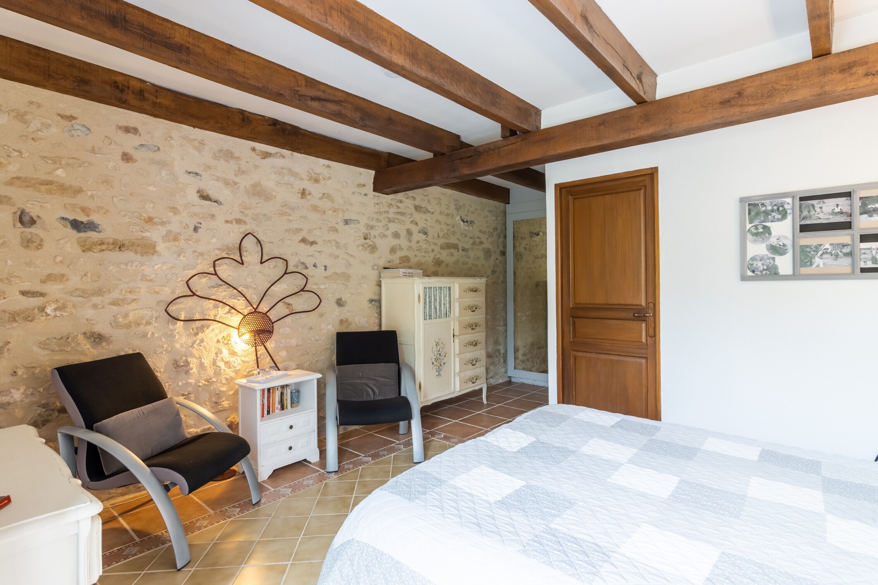 Une chambre avec un mur en pierre, des poutres en bois, un lit, une chaise, une petite table et un meuble.