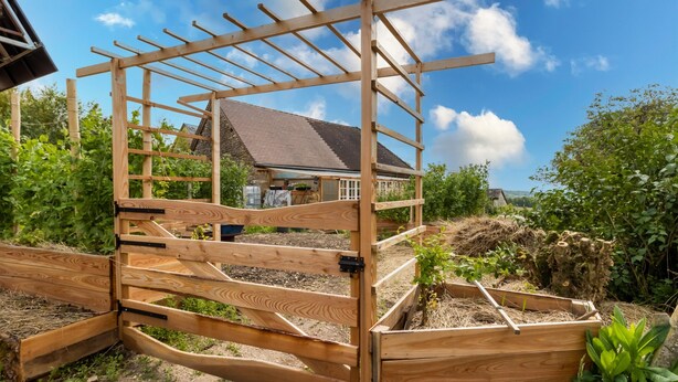 Una verja de madera con estructura de pérgola, un cantero con plantas y una casa al fondo.