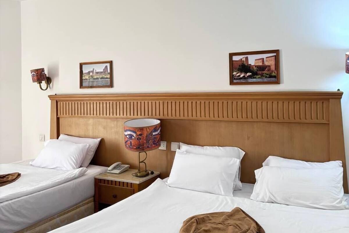 Una camera d'albergo con due letti singoli, una testiera in legno, un comodino con una lampada e due quadri appesi al muro.