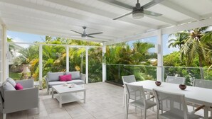 A covered outdoor patio with a white ceiling, ceiling fans, a white table, white chairs, and a grey sofa with pink cushions.