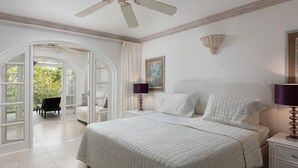 A bedroom with a large bed, a nightstand, a lamp, a painting, and a view through an open door to a balcony with plants.