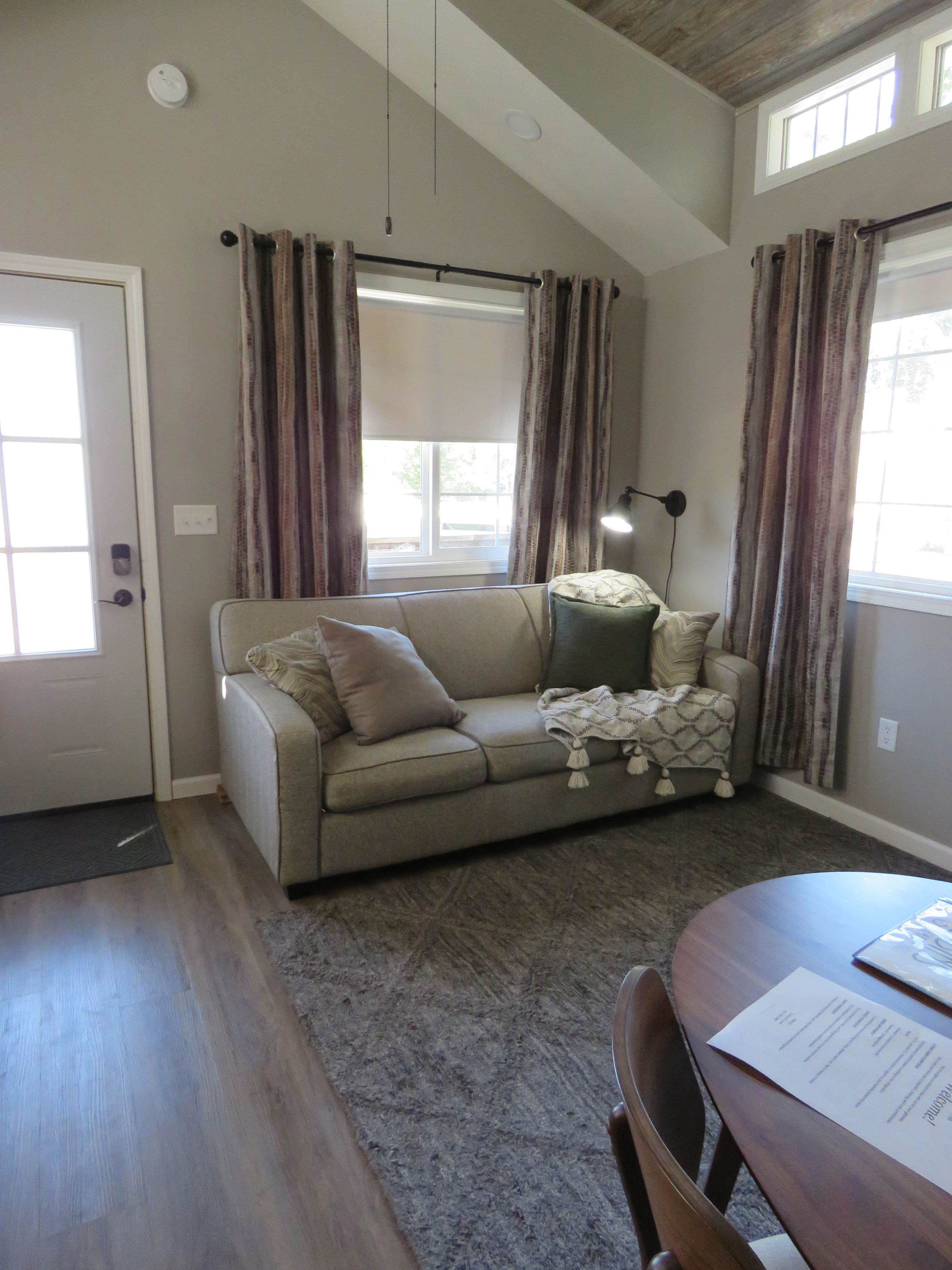 A living room with a sofa, a table, and a lamp. There is a window with curtains and a door.