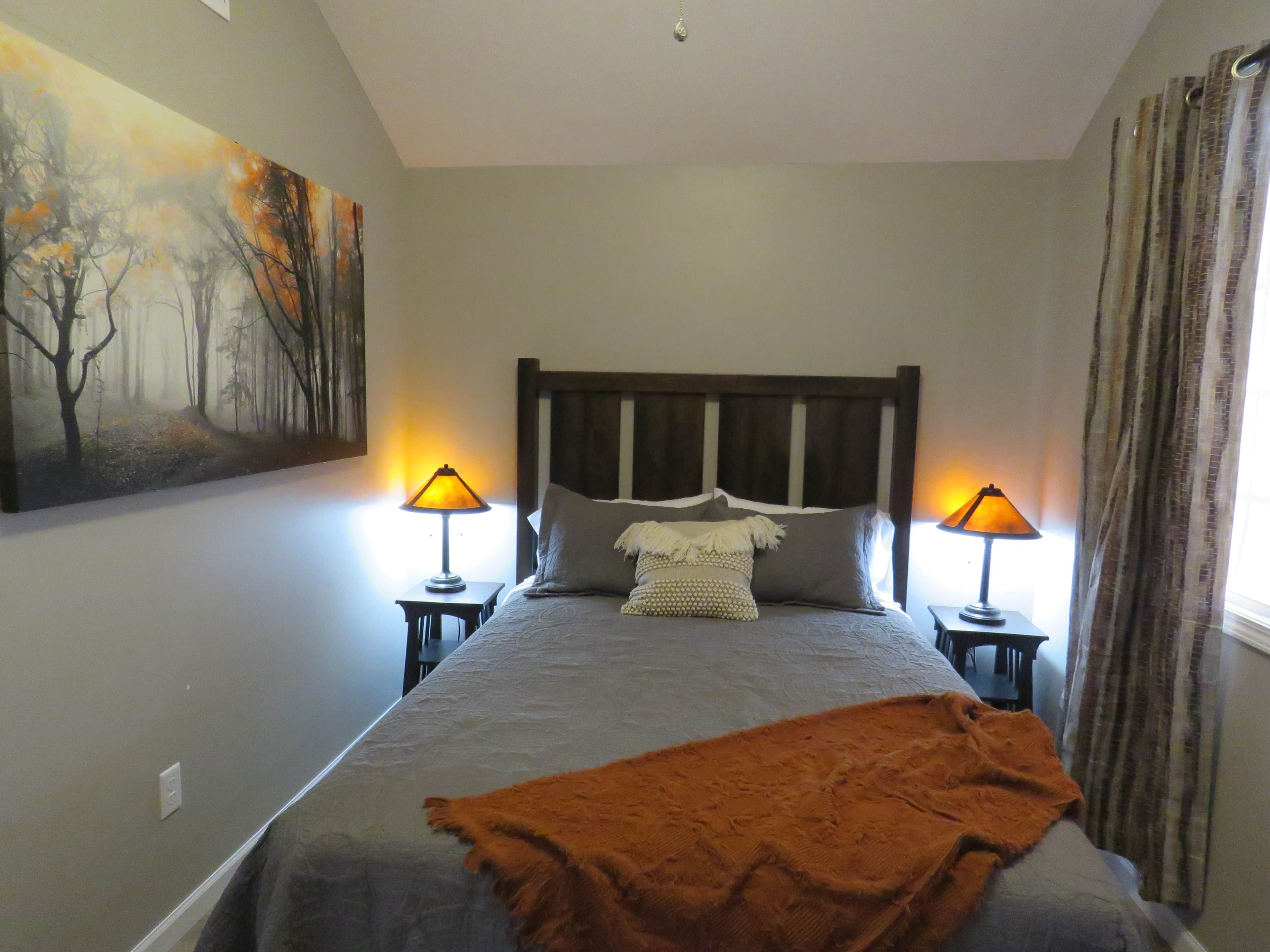 A bedroom with a bed, two bedside lamps, a painting on the wall, and a window with curtains.