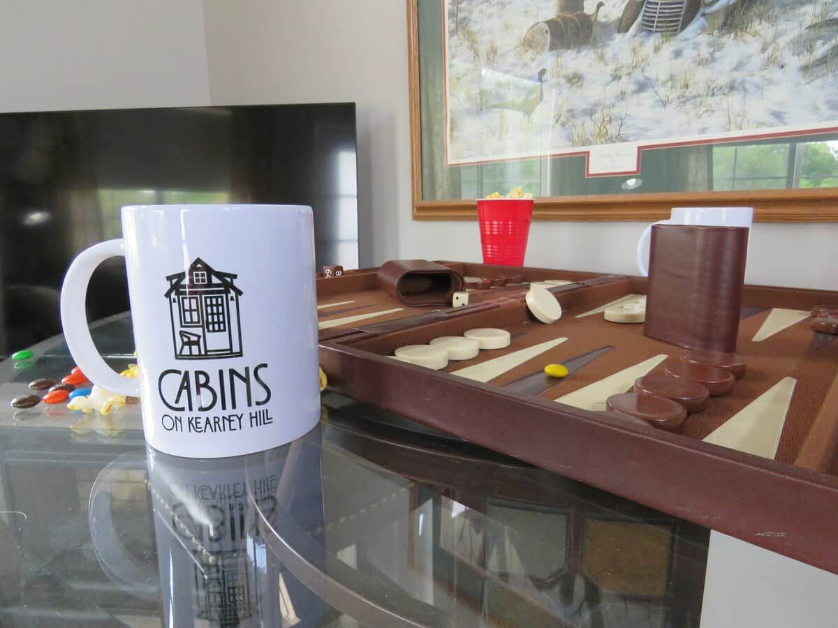 Una taza de café con el logotipo de una cabaña sobre una mesa con un juego de mesa de cuadros, un vaso rojo y un televisor al fondo.