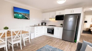 A modern kitchen with white cabinets, a stainless steel refrigerator, a microwave, and an oven. There is a dining table with white chairs and a blue patterned rug. - 2B at Swansea - cosy cottage, outdoor pet friendly (Swansea)