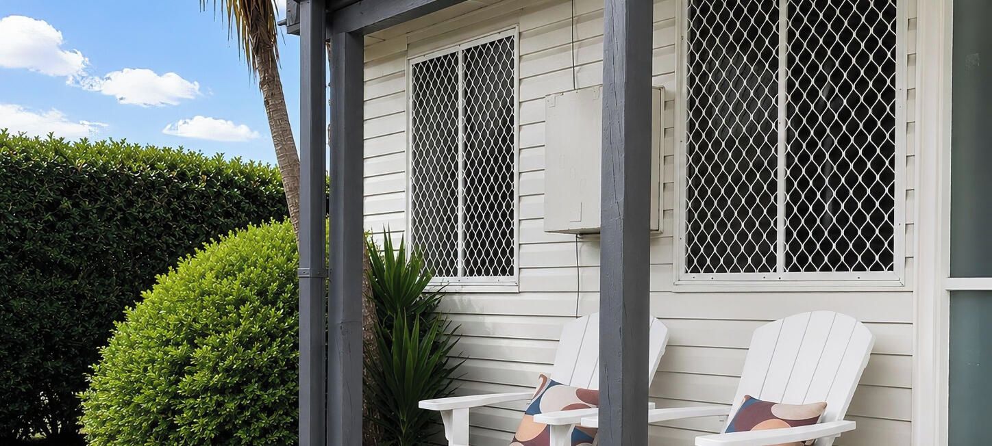 A house with two windows with metal grilles, a white exterior, and two white Adirondack chairs with cushions.