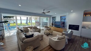 A spacious living room with a large sofa, armchairs, and a coffee table. There is a television, a kitchen area with a microwave, and a dining table. The room has large windows offering a view of a pool and greenery. - Lake Sabai - Waterfront and private pool (Nords Wharf)