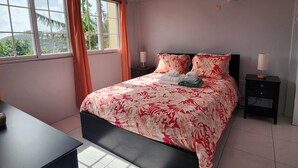 A bedroom with a bed, two bedside tables, an air conditioner, a window with curtains, and a view of greenery outside.