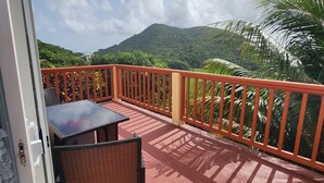 A balcony with a table and chairs, a red railing, and a view of green hills and a clear sky.