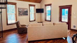 Ein geräumiges Wohnzimmer mit Holzbalkendecke, großen Fenstern, einem Flachbildfernseher und einem gemütlichen Sofa.