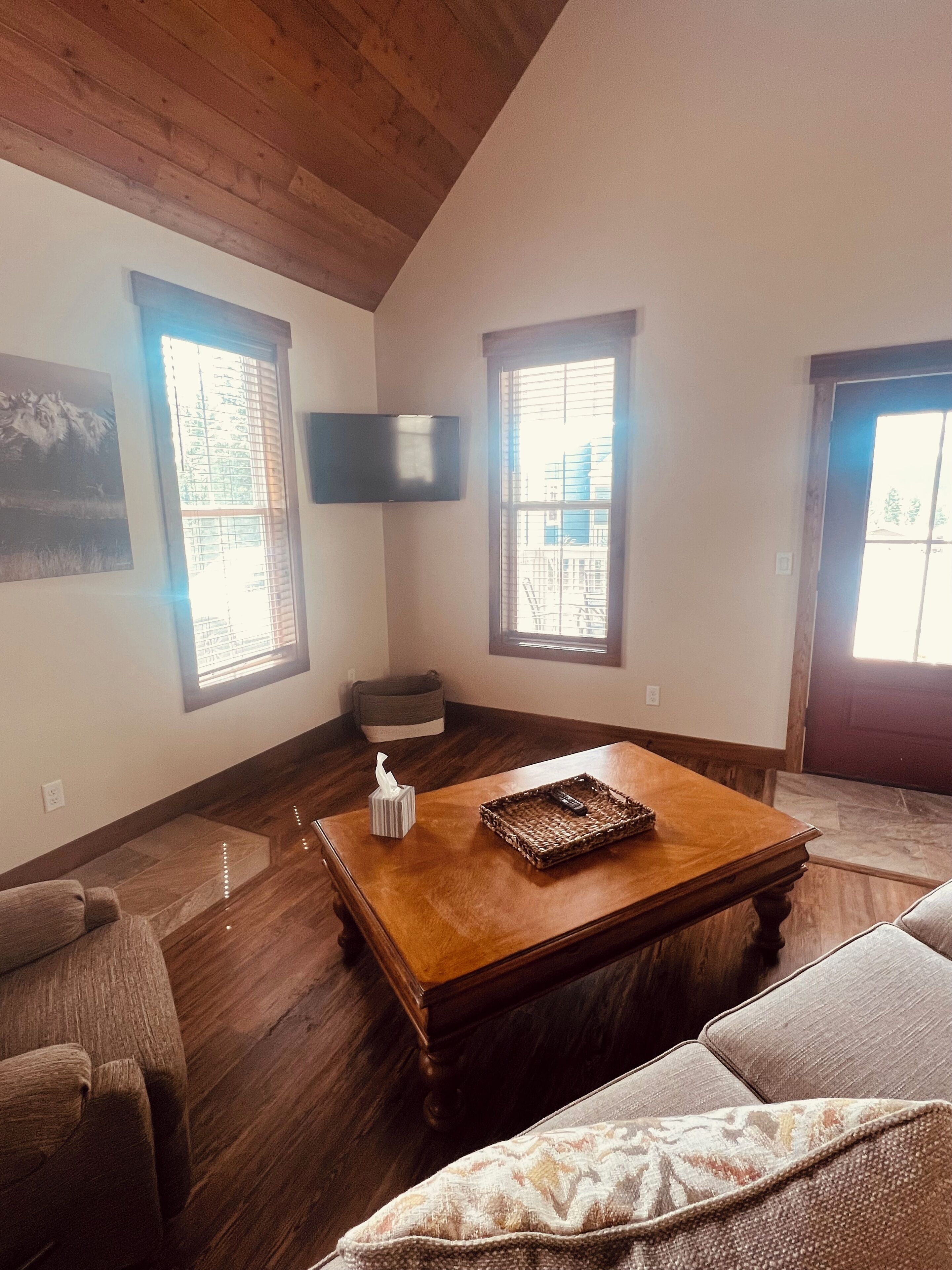 A living room with a wooden coffee table, two windows with blinds, a flat-screen TV mounted on the wall, and a couch.