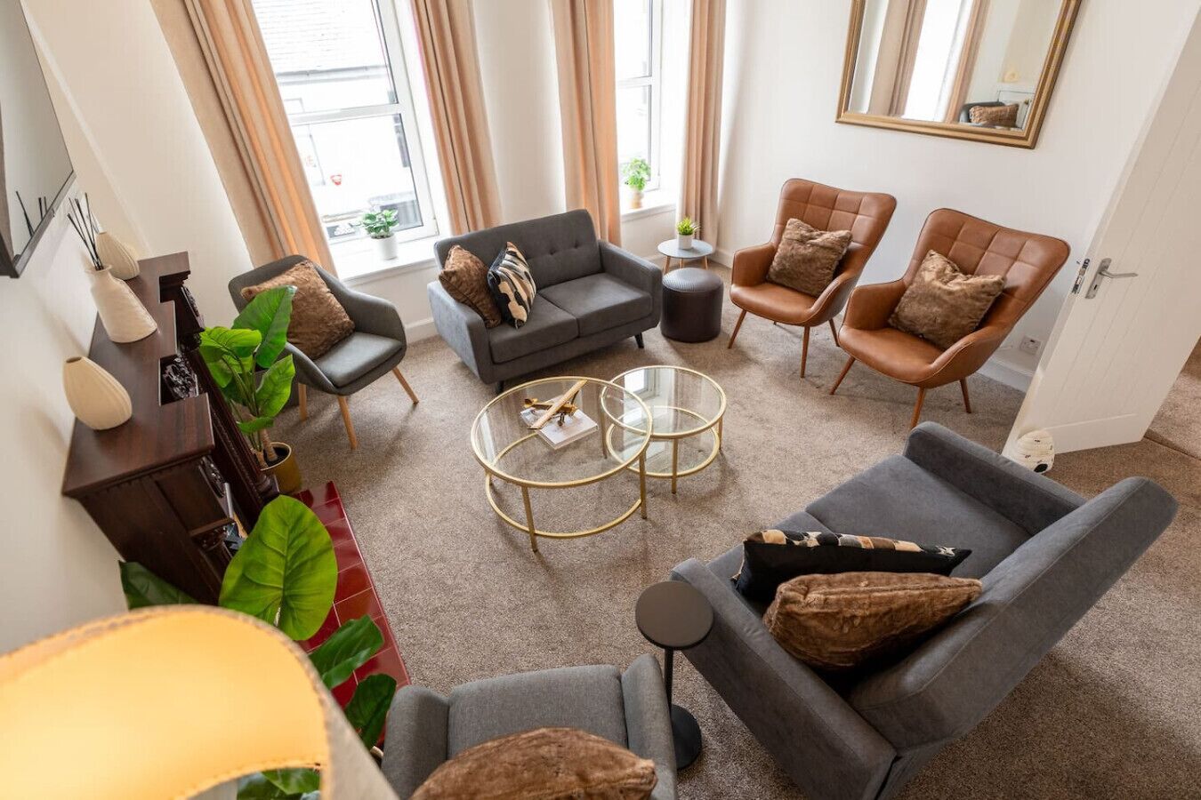 A modern living room with a grey sofa, two armchairs, a coffee table, and a small side table. There are potted plants and a large mirror on the walls.