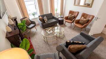 A modern living room with a grey sofa, two armchairs, a coffee table, and a small side table. There are potted plants and a large mirror on the walls.