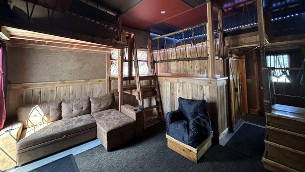 A cozy cabin interior with a sofa, a cushioned bench, a wooden ladder, and a loft area with string lights.