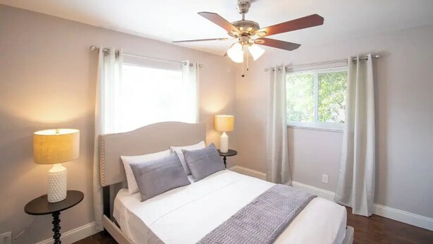 Une chambre à coucher avec un lit, deux lampes de chevet, un ventilateur de plafond et une fenêtre avec des rideaux.