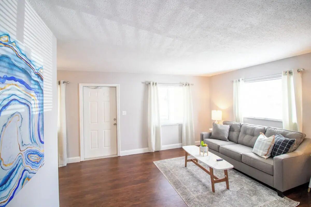 Una sala de estar con un sofá gris, una mesa de centro blanca y un tapete estampado. Hay una puerta y una ventana con cortinas.