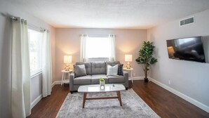 Un salon avec un canapé gris, deux tables d’appoint avec des lampes, une table basse, un téléviseur fixé au mur et une fenêtre avec des rideaux.