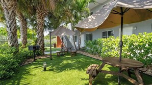 Un espace extérieur aménagé avec soin, comprenant une terrasse, une table en bois et des bancs, le tout entouré d’une végétation luxuriante et de palmiers.