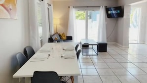 Un comedor con una mesa larga dispuesta para cuatro, un ventilador de techo, un televisor montado en la pared y vistas a través de la puerta de cristal.