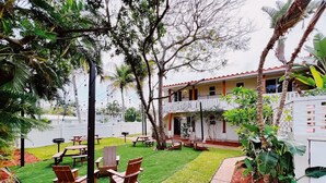 A patio area with wooden chairs and tables, surrounded by lush greenery and a white building with a red roof.