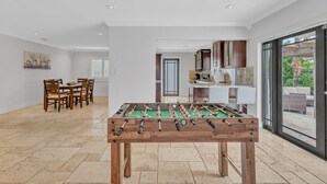 Une pièce spacieuse équipée d’un baby-foot, d’un coin repas avec des chaises en bois et d’une cuisine avec des appareils modernes.