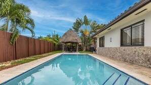 Une piscine avec une structure dotée d’un toit de chaume en arrière-plan, entourée de palmiers et d’une clôture en bois.