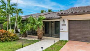 Une maison d’habitation avec un toit en tuiles, un portail de garage et un jardin bien entretenu, agrémenté de palmiers et d’arbustes.