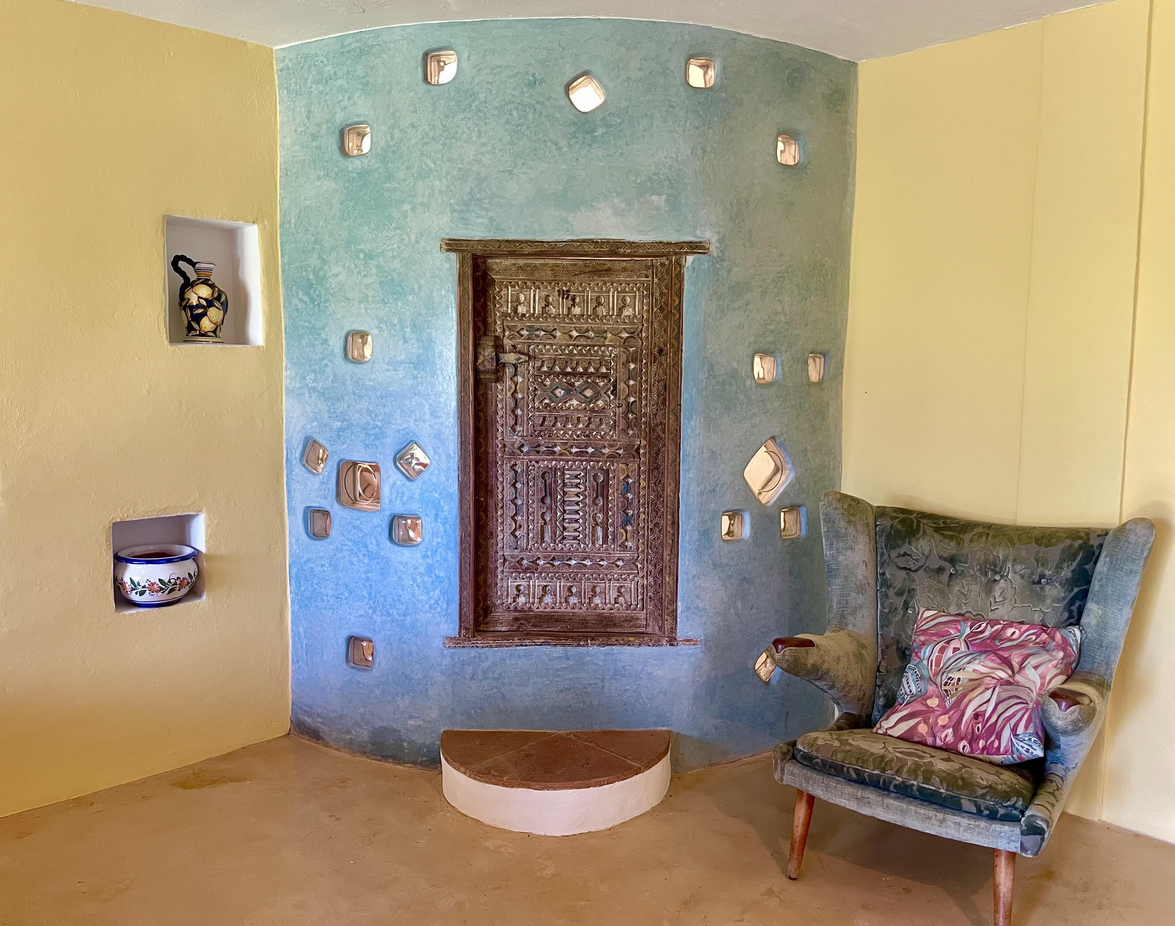 Una stanza con una parete blu decorata, una porta in legno con intagli elaborati, un piccolo ripiano con oggetti decorativi e una poltrona con motivi fantasiosi.