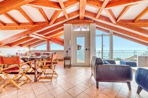 Een ruime eethoek met houten stoelen en een tafel, een balkon met blauwe stoelen en uitzicht op zee.