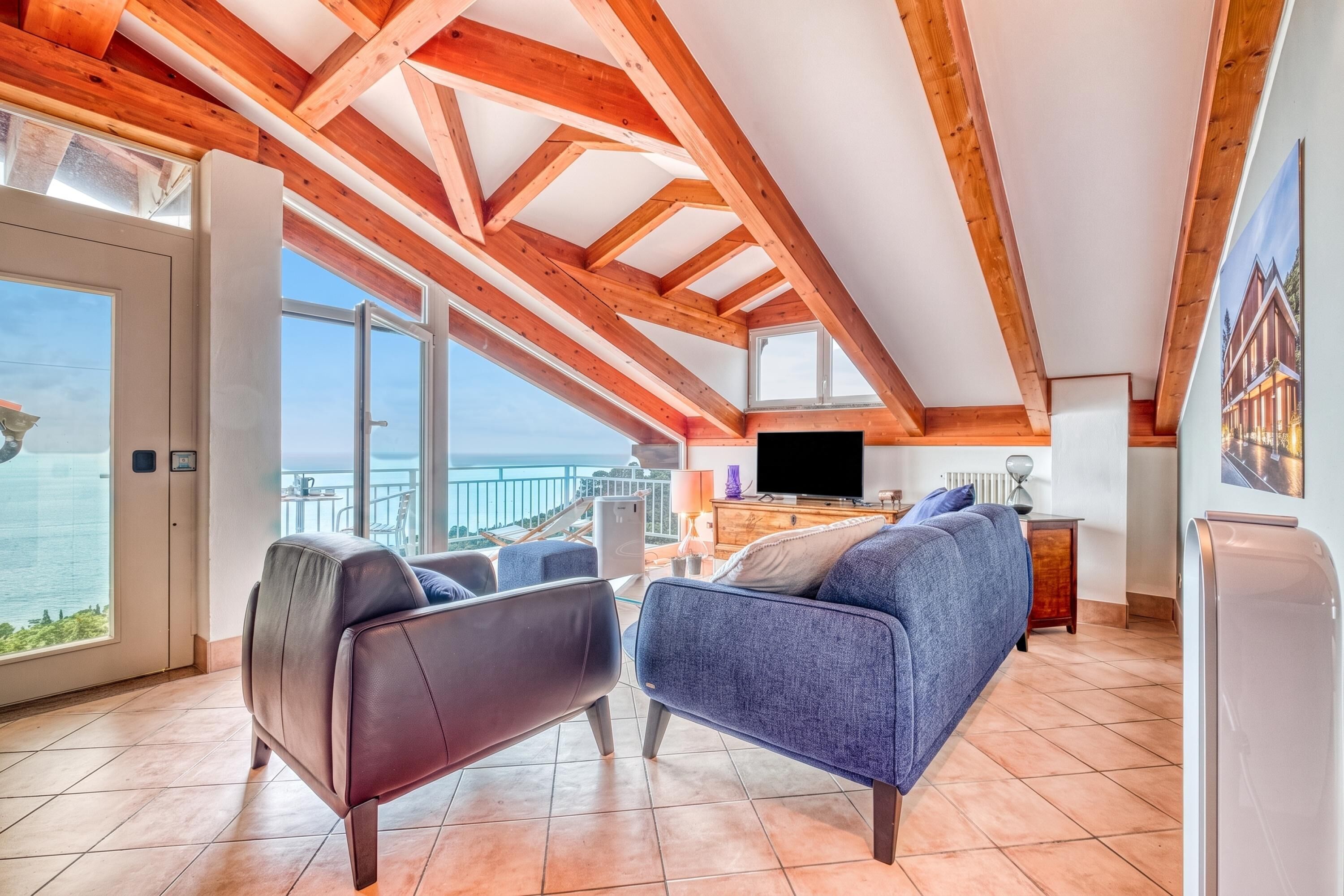 A spacious living room with a large window overlooking a body of water, wooden beams on the ceiling, a flat-screen TV, and a sofa.