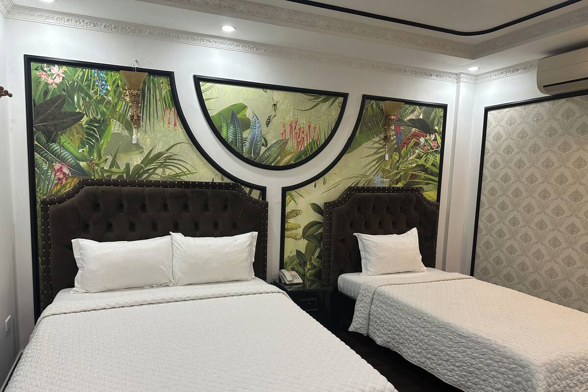 A hotel room with two beds, each with a tufted headboard and white bedding. The walls are decorated with tropical wallpaper, and there is a patterned wallpaper on one side.